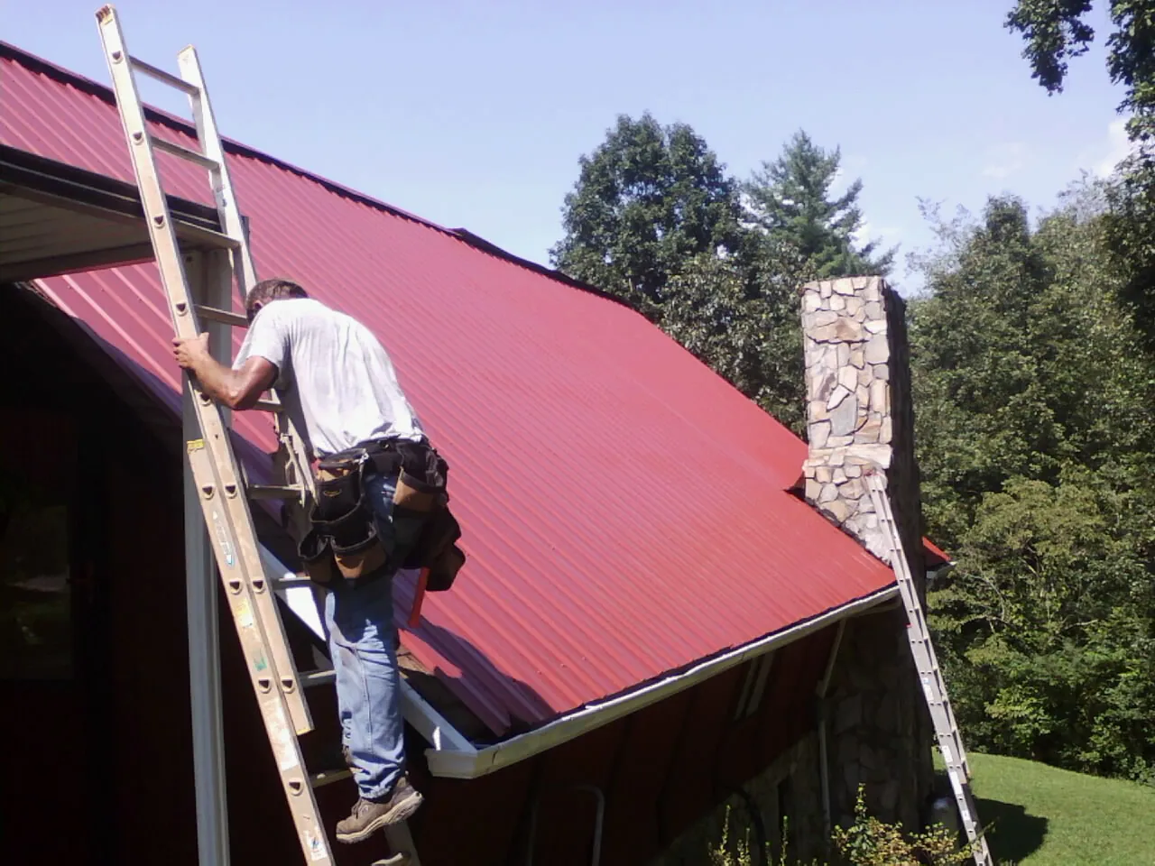 Roofer on a ladder beside a red metal roof and stone chimney — Soffit & Fascia Repair by Roofing Brothers in Stockdale